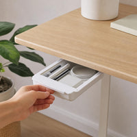 Une main ouvre le tiroir HomeTuck blanc collé sous un bureau en bois clair : stylos et petite boîte ronde à l'intérieur